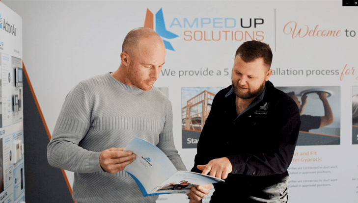 Two men reviewing a brochure together in front of a display with the "Amped Up Solutions" logo