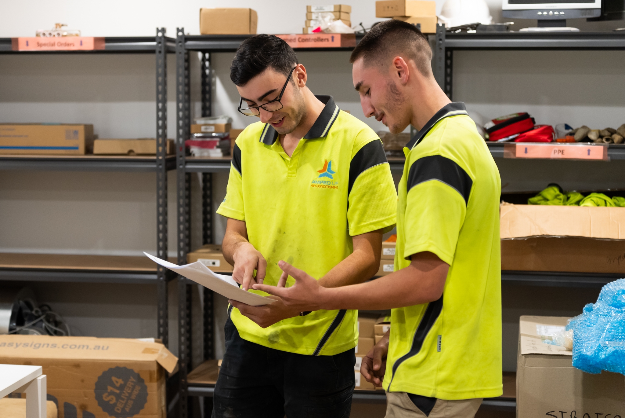 Two men in yellow high-visibility shirts review paperwork together in a warehouse or storage room with shelves and boxes in the background.