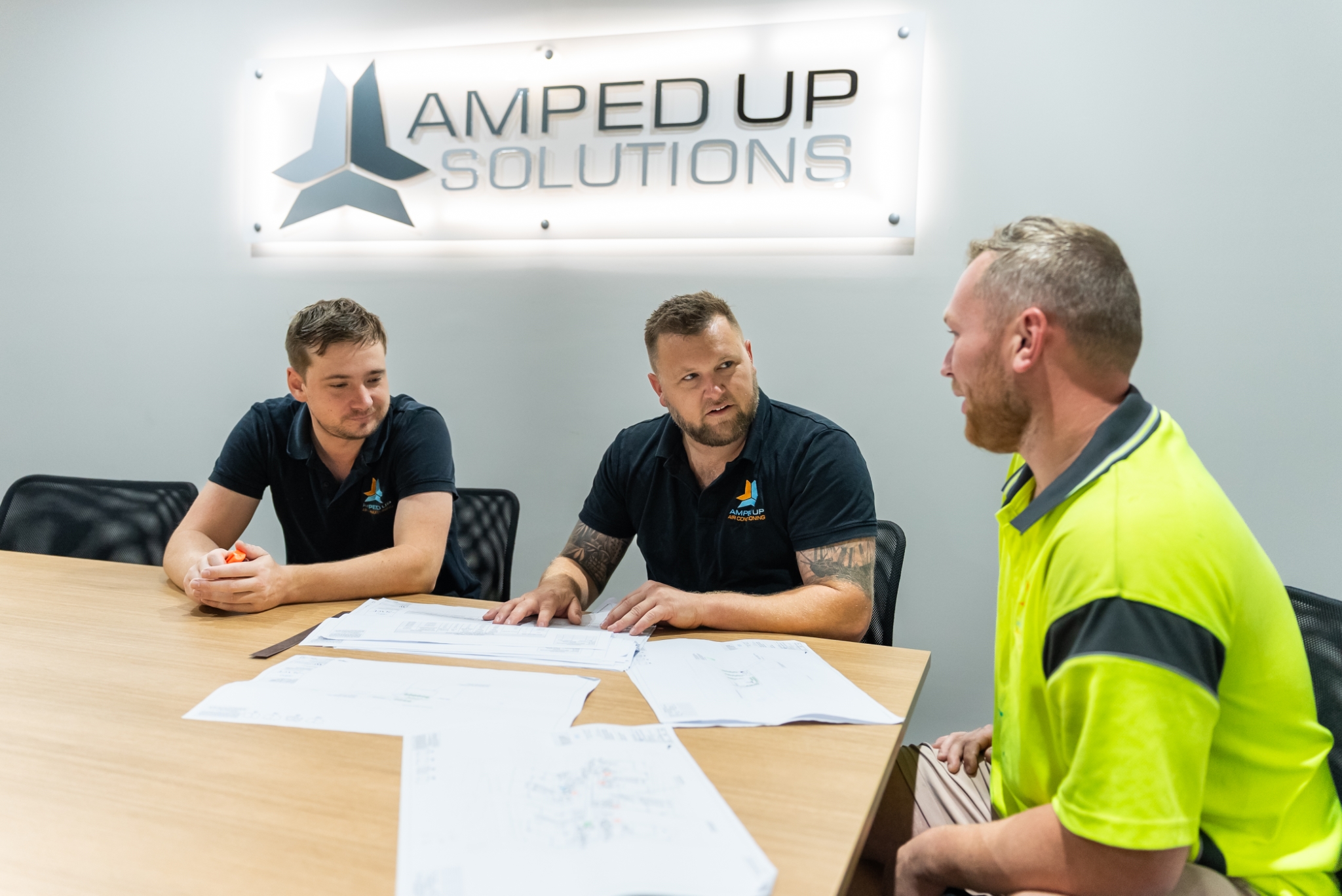 Three men sit at a conference table discussing documents under a sign that reads "Amped Up Solutions.
