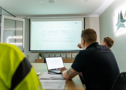 Three people in an office meeting room review a floor plan projected on a screen; one person points at the screen, while another uses a laptop.