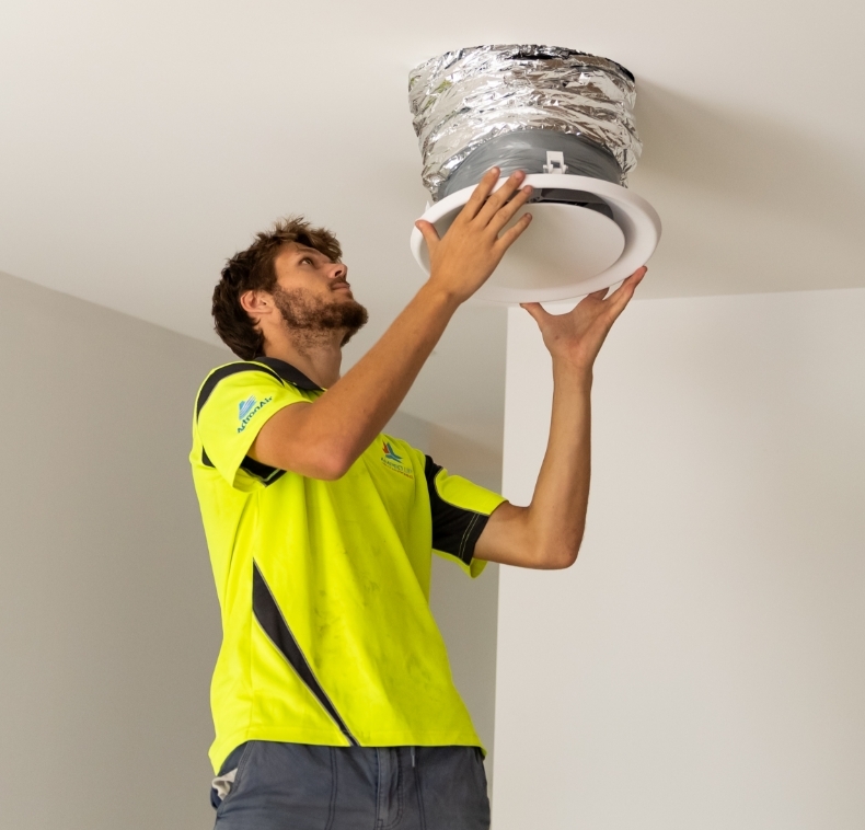 A worker in a yellow shirt is installing or repairing a round ceiling air vent with exposed ductwork.