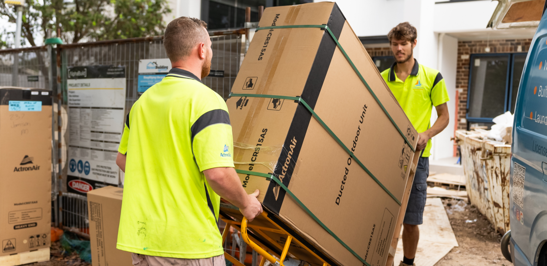 Two workers in yellow safety shirts using a hand truck to move a large, boxed ducted outdoor unit