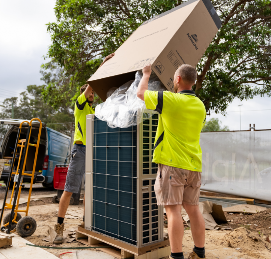 Two workers in yellow shirts remove packaging from a large air conditioning unit outdoors