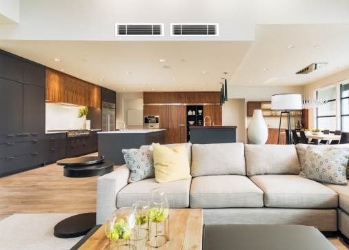 Modern open-concept living room and kitchen with neutral decor, light sofa, wooden accents, and bar stools at a kitchen island.