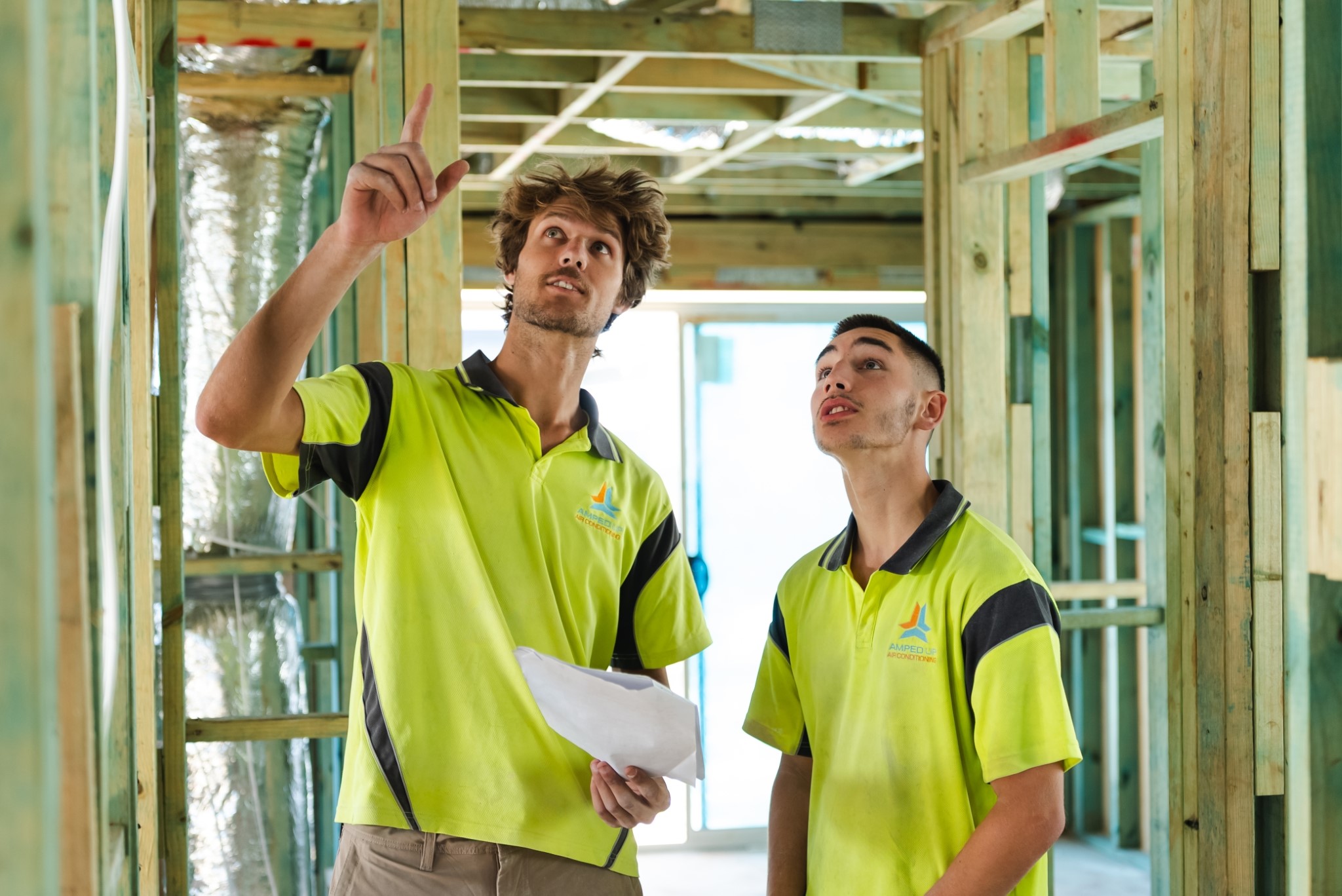 Two construction workers in yellow shirts inspecting the wooden framing of a building