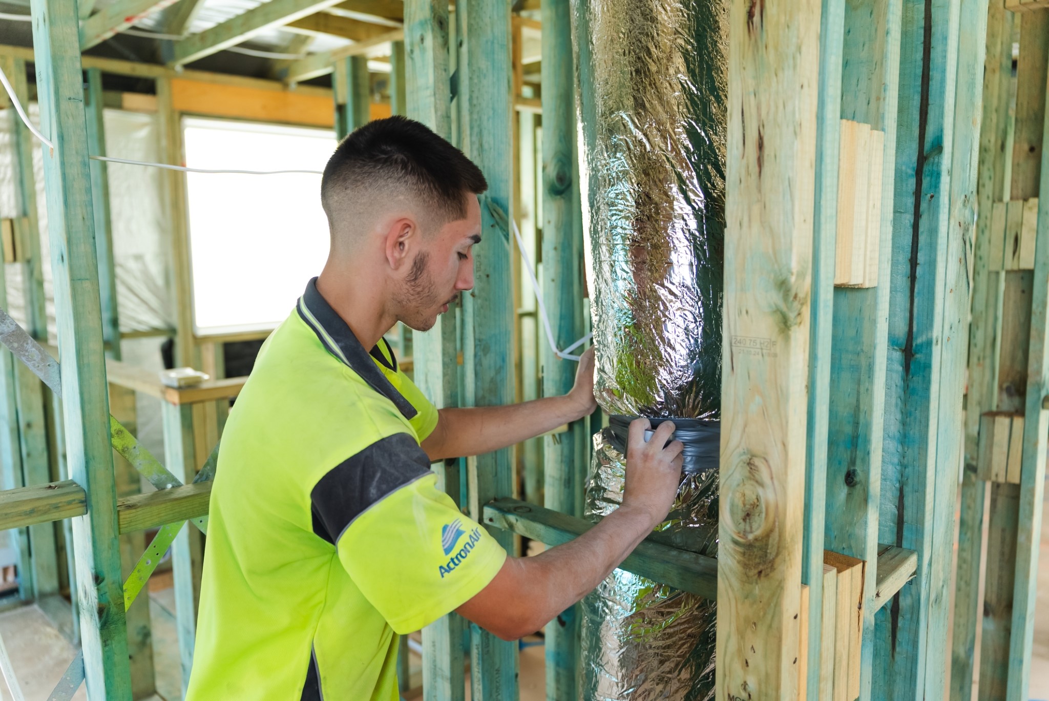 A worker in a high-visibility shirt applies tape to a foil-wrapped duct inside the wooden frame of a building under construction.