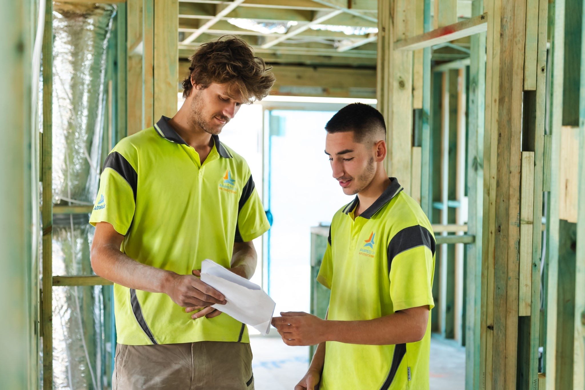 Two construction workers in yellow shirts review documents together inside a building under construction with exposed wooden framing.