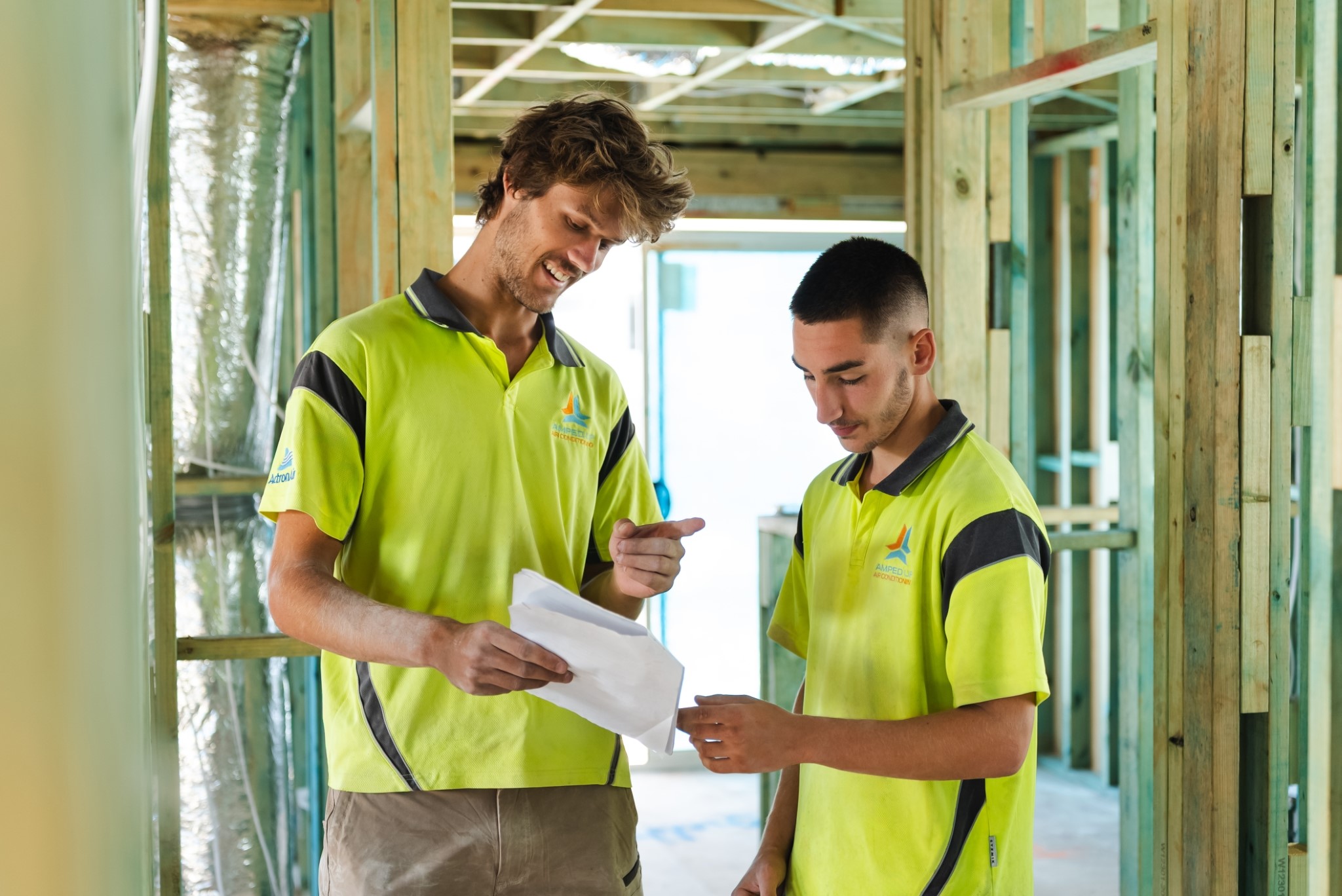 Two construction workers in yellow shirts reviewing a paper together inside a building
