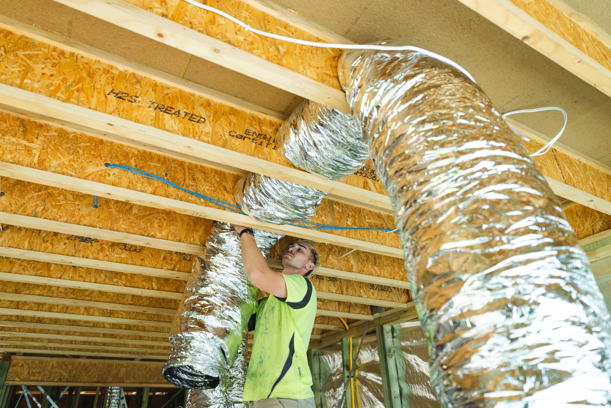 A person installs flexible HVAC ducts and electrical wiring in the ceiling framework of a building under construction.