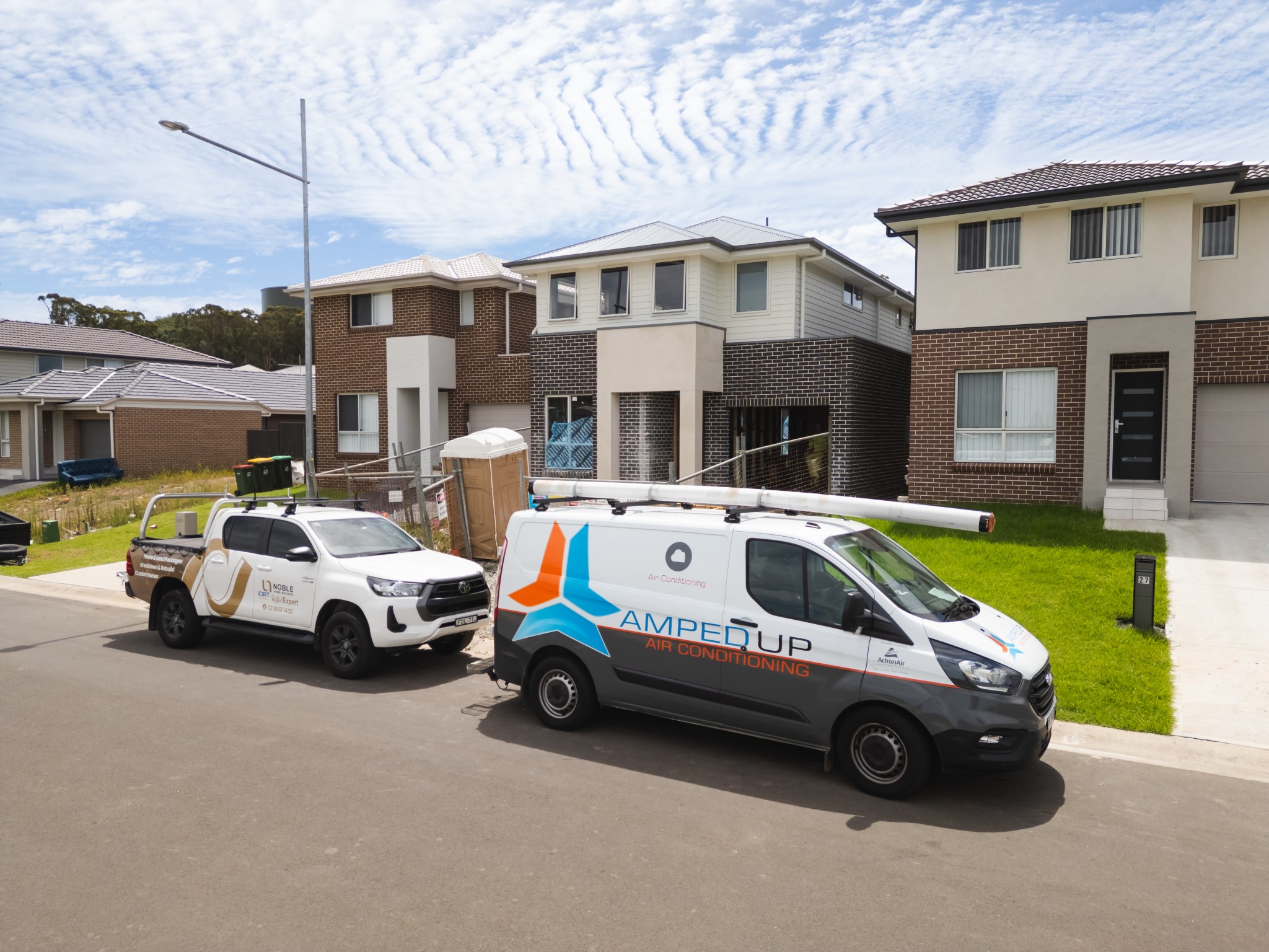 Two service vehicles, one labeled "Amped Up Air Conditioning," are parked on the street in front of modern suburban houses during the day.
