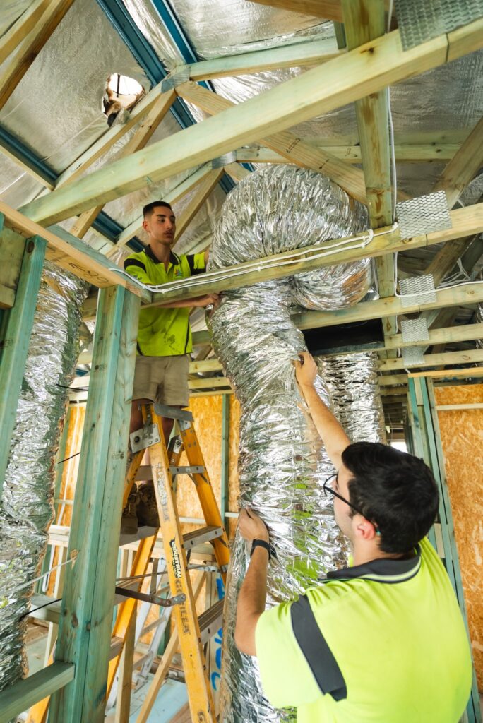 Two workers in high-visibility shirts install flexible ductwork in the ceiling of a building