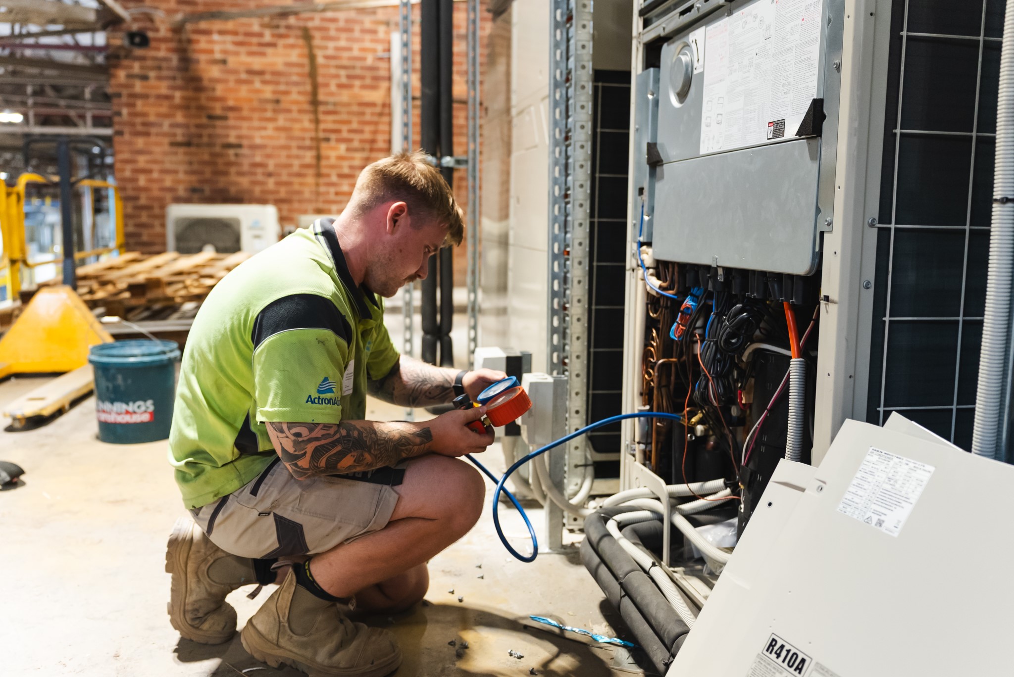 A technician in a green shirt and shorts kneels to inspect and work on electrical components in an open utility panel indoors.