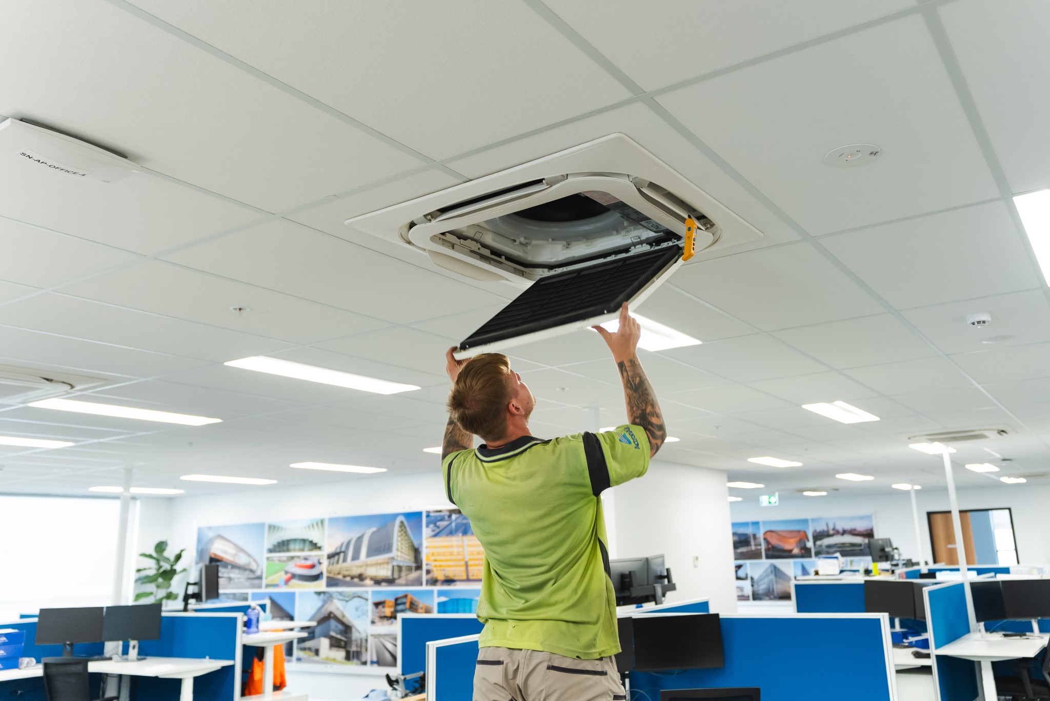 A person in a green shirt removes a filter from a ceiling-mounted air conditioning unit in a modern office.