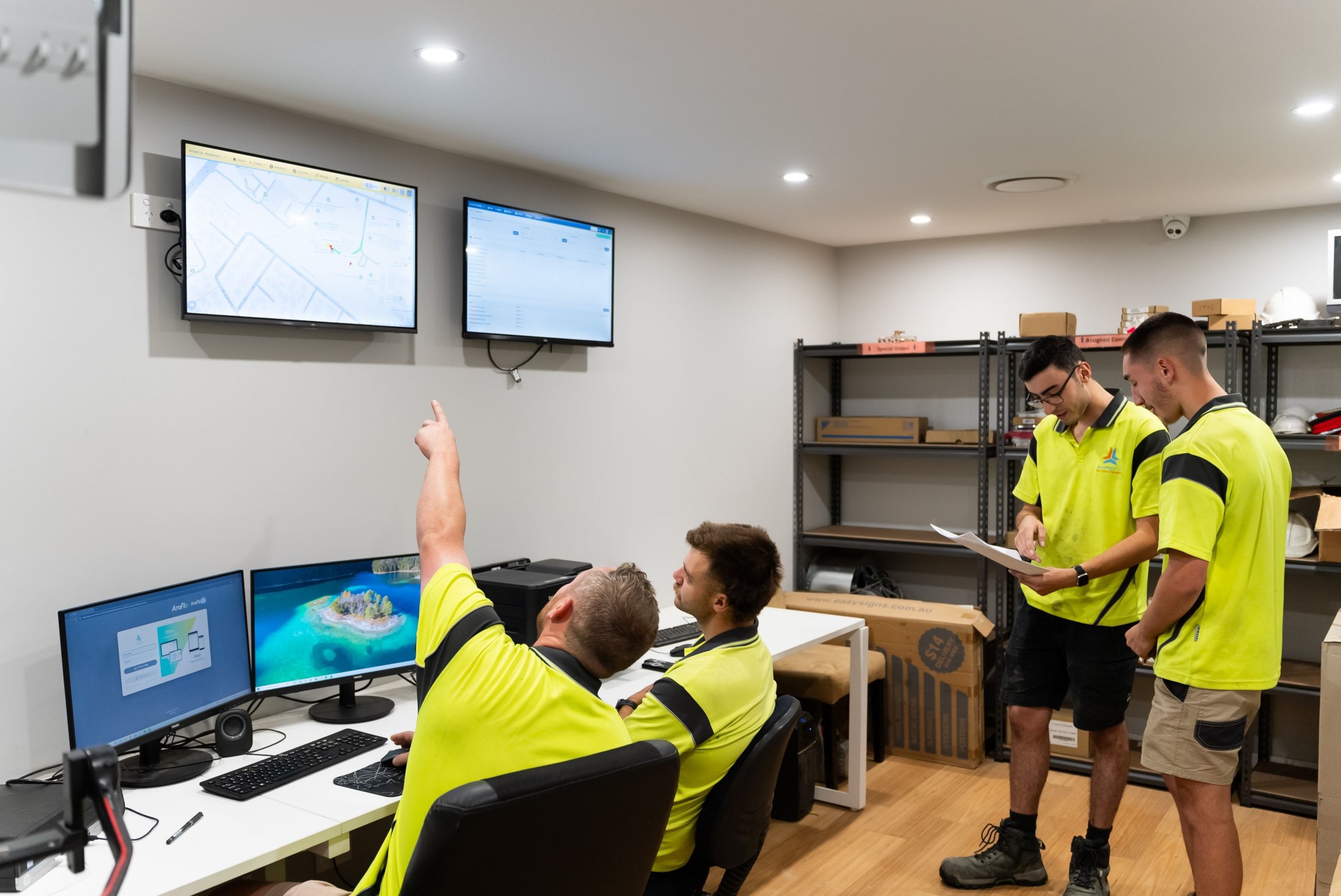 Four men in yellow uniforms work in an office with multiple monitors and shelves; one points at a screen displaying a map while others review documents.