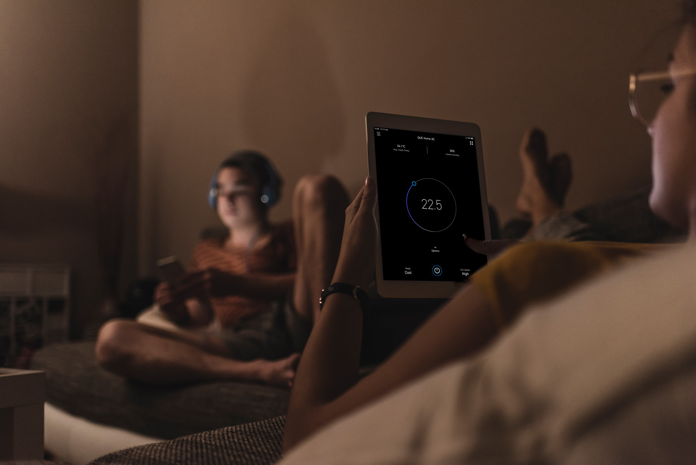 Person on a couch holds a tablet displaying a smart home control screen, while another person with headphones sits in the background using a smartphone.